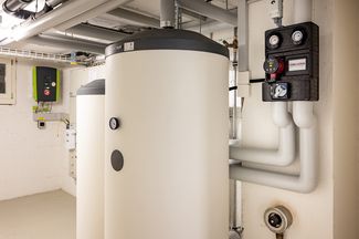 Das Bild zeigt einen Technikraum mit grossen, zylindrischen Warmwasserspeichern in hellen Farben. Rechts daneben befindet sich eine Pumpen- und Regelungseinheit mit zwei Druckmanometern, einer elektronischen Pumpe und mehreren angeschlossenen Leitungen. Die Rohrleitungen sind sauber verlegt und führen sowohl zu den Speichern als auch in die Wand. Die Umgebung wirkt ordentlich und technisch modern ausgestattet.